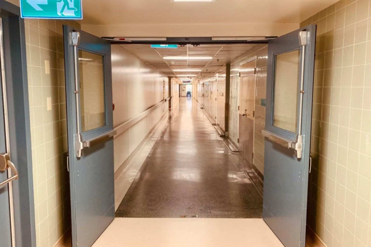 Perspectief van een lange verlichte ziekenhuisgang, omlijst door glazen branddeuren met paniekstangen